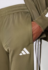 Olívazöld tréningnadrág sima anyagból, fehér Adidas logóval és fehér oldalsávval. Rugalmas, ráncolt derékpánt.