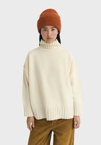 Junge Person in einem cremfarbenen Rollkragenpullover, einer brennen-orangen Strickmütze und braunen Cordhosen, die vor einem schlichten Hintergrund steht.