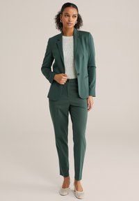 Sieviete stāv, ģērbusies tumši zaļā pieguļošā žaketē un biksēs, ar gaišu trikotāžas topu un krēmkrāsas augstpapēžu kurpēm, neitrāls fons.