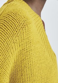 Pull jaune tricoté avec un col en V et un motif texturé. Le tissu semble doux avec un tissage lâche et des accents côtelés le long de l'encolure.