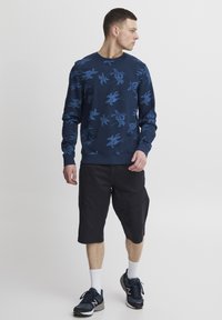 Marineblå genser med blått blomsterdesign, kombinert med svarte shorts. Modellen har på seg hvite sokker og mørke joggesko, stående mot en nøytral bakgrunn.