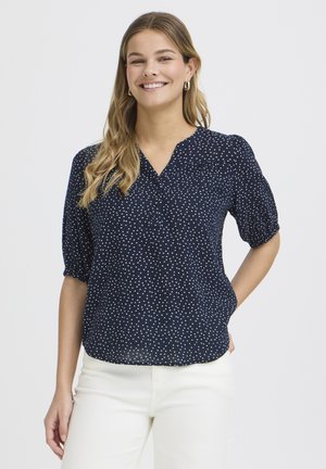 Kobieta uśmiechająca się, ubrana w granatową bluzkę w białe kropki i białe spodnie, stojąca z jedną ręką za plecami.