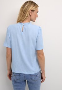Top azzurro chiaro a maniche corte in tessuto testurizzato, con scollo arricciato e una piccola apertura a chiave sulla schiena con chiusura a bottone. Design semplice, vestibilità comoda.