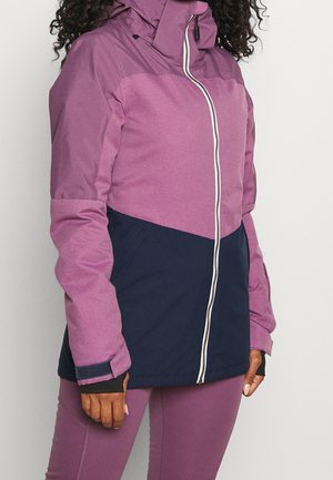 Kobieta w dwukolorowej fioletowo-granatowej rozpinanej kurtce z wysokim kołnierzem, otworami na kciuki w rękawach i dopasowanych legginsach.