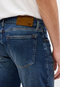 Jeans in denim blu scuro con una texture liscia, caratterizzati da una toppa in pelle marrone sulla vita e cuciture arancioni a contrasto.