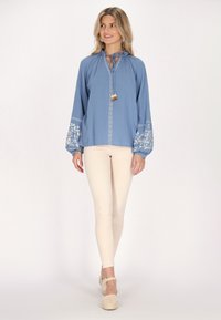 Blusa azzurro chiaro con collo rotondo, dettaglio con fiocco e maniche lunghe ricamate. Indossata con pantaloni skinny beige e scarpe espadrilles.