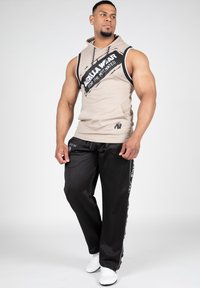 Beige hoodie zonder mouwen met zwarte grafische print; zwarte, losse broek met zijtekst; witte sneakers. Glad materiaal, casual ontwerp.