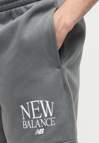 Χέρι στην τσέπη που φορά γκρι φόρμα New Balance με λευκό λογότυπο στο μηρό, σε απλό φόντο.