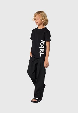 Enfant blond debout, portant un T-shirt noir avec "KARL" imprimé verticalement, un pantalon noir ample et des sandales noires avec un texte blanc.