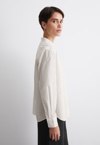 Chemise blanche à manches longues avec des pois noirs, coupe ajustée, poignets boutonnés et ourlet arrondi. Tissu doux au toucher lisse.