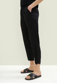 Zwarte broeken met een elastische tailleband en zijzakken, met een taps toelopend ontwerp en subtiele zijplooien. Gecombineerd met zwarte instapsandalen.
