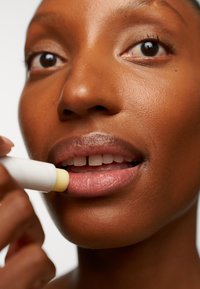 Nahaufnahme der Lippen einer Person, die einen weißen Lippenbalsam mit einer gelben Spitze aufträgt. Die Lippen sind hellrosa, die Haut ist glatt und tiefbraun.