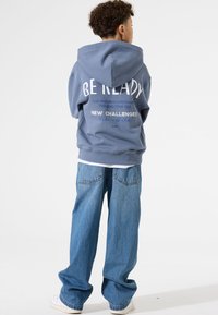 Hellblaue Kapuzenjacke mit weißem Text auf dem Rücken, kombiniert mit lockeren blauen Jeans und weißen Sneakern.