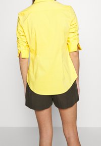 Gul skjorta med skräddarsydd passform, med samlade ärmar och krökt nederkant. Lättviktig tyg med en mjuk textur. Kombinerad med bruna shorts.