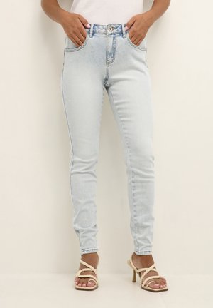 Kvinde iført lyseblå skinny jeans og beige hælede sandaler med flettede remme, hænderne i lommerne, stående mod en ensfarvet baggrund.