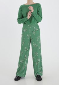 Grön stickad tröja med bred halsringning, kombinerad med gröna byxor med blommönster och vida ben, som har vita blommor och mörka grenar.