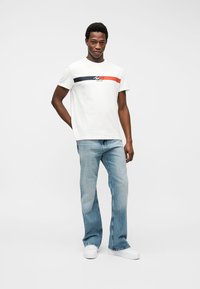 Άνδρας που στέκεται φορώντας λευκό T-shirt με μπλε και κόκκινες ρίγες, ανοιχτό μπλε τζιν και λευκά αθλητικά παπούτσια σε ένα απλό φωτεινό φόντο.