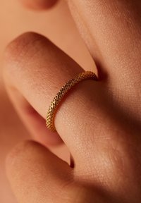 Goldring mit einem strukturierten, geflochtenen Muster, der an einem Finger getragen wird. Die Oberfläche ist glatt und glänzend, was die komplexen Designdetails zur Geltung bringt.