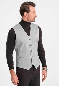 Grau karierter Weste mit tiefem V-Ausschnitt, fünf braunen Knöpfen und zwei Fronttaschen über einem dunklen Rollkragenpullover. Schlankes Design.
