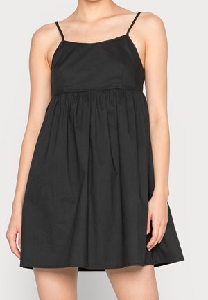 Nainen pukeutuneena hihattomaan mustaan mekkoon, jossa ohuet hihnat ja kerätty vyötärö, seisoo yksinkertaista vaaleaa taustaa vasten.