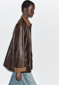 Veste en cuir marron avec un col beige, présentant une silhouette décontractée et de grandes poches. Portée avec un jean en denim bleu clair.