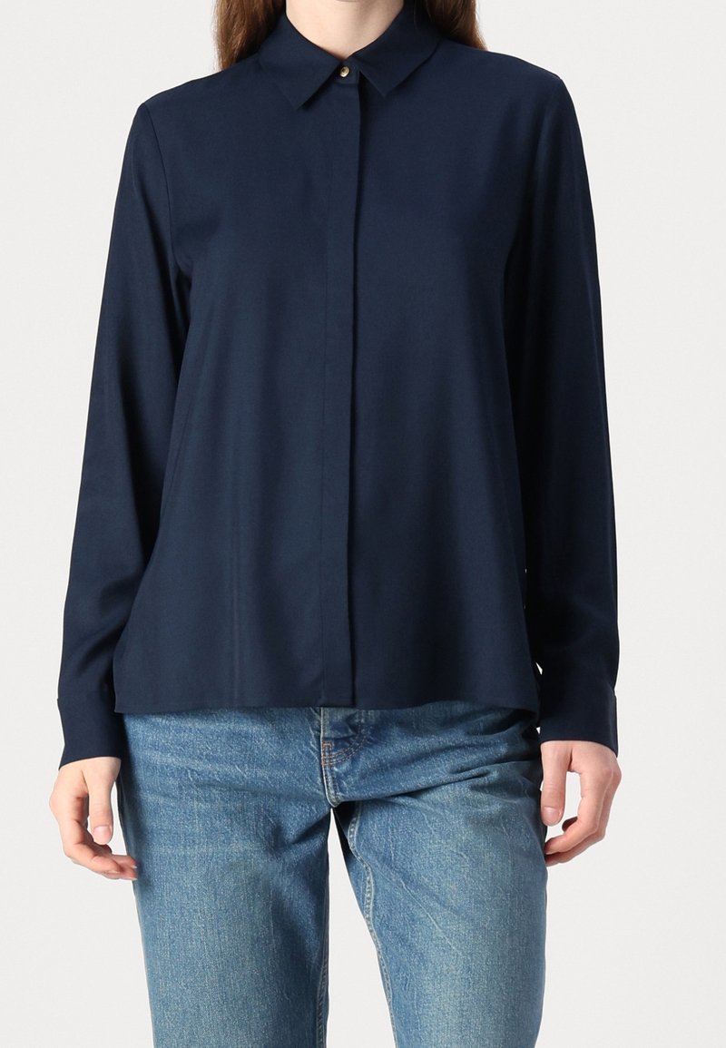 Personne portant une chemise à manches longues bleu marine avec col et boutons cachés et un jean bleu, debout devant un fond uni clair.