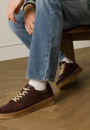 Bordeaux suede sneakers met lichtbeige veters, rubberen zool en geperforeerde zijdetails, gedragen met blauwe spijkerbroeken en witte sokken.