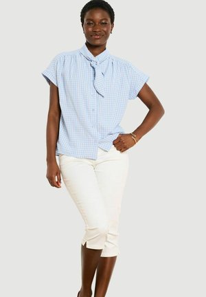 Kobieta stoi pewnie, nosząc niebieską kraciastą bezrękawnikową bluzkę z kokardą na szyi oraz białe krótkie spodnie na jednolitym tle.