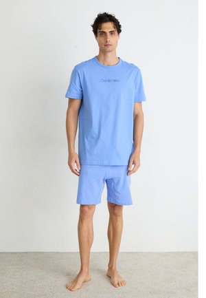 Camiseta de algodón azul claro con cuello redondo y logo de "Calvin Klein", combinada con pantalones cortos azules claros a juego. Diseño simple y casual.