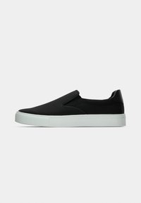 Chaussures noires sans lacets avec une tige en tissu lisse, des panneaux latéraux élastiques et une semelle en caoutchouc blanche. Design minimaliste sans motifs visibles.
