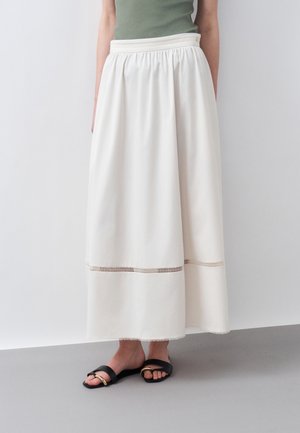 Osoba nosiaca bielu sukňu s vysokým pásom a dĺžkou po členky, s horizontálnym výrezom nad lemom a čierne šľapky na slip-on.