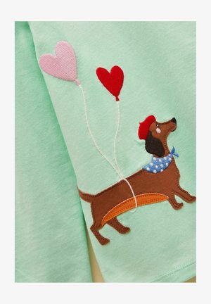 Brauner Dackel mit roter Baskenmütze und blauem, gepunktetem Schal, befestigt an rosa und roten herzförmigen Luftballons auf hellgrünem Stoff.