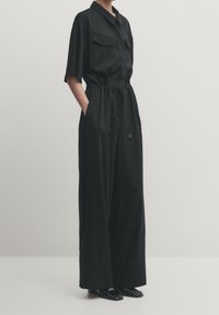 Zwart jumpsuit met korte mouwen, twee borstzakken, taillekoord, wijde benen en een gladde textuur. Draagt met glanzende zwarte schoenen.