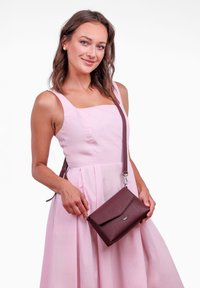 Bordo křížová taška vyrobená z texturované umělé kůže, s plochým klopovým zavíráním a stříbrným akcentem. Nosená modelem v růžových šatech.