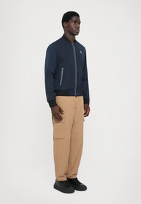 Casaco bomber azul-marinho com fecho de correr à frente, piping branco e punhos canelados, combinado com calças cargo de tom claro e sapatos pretos.