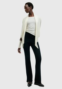 Cremfarbener Strickcardigan, der über einem enganliegenden weißen Oberteil und schwarzen Schlaghosen getragen wird. Strukturierter Stoff mit schmaler Rippung und subtilen Details auf der Kleidung.