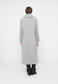 Abrigo largo gris con cuello de piel sintética, de corte recto y con detalles mínimos. Textura suave, que llega a mitad de la pantorrilla. Llevado sobre pantalones oscuros y ajustados.