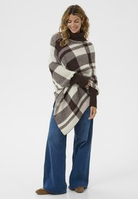 Rudas ir kreminis languotas pončas su aukštu apykakle ir plačiomis rankovėmis, krentantis ant mėlynų plačių kojų džinsų. Lygi, tekstūruota medžiaga.