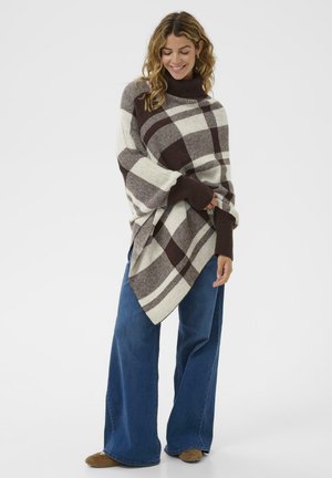 Hnědo-krémový kostkovaný pončo s vysokým límcem a širokými rukávy, přehozené přes modré široké kalhoty. Hladká, texturovaná tkanina.