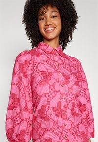 Blusa floral rosa com bordado texturizado, fecho de botão e colarinho clássico. Apresenta mangas bufantes e padrões florais variados.