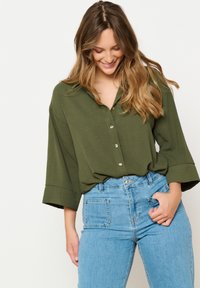 Olive green, loose-fit button-up shirt with rolled sleeves. Paired with light blue high-waisted jeans featuring front and back pockets.