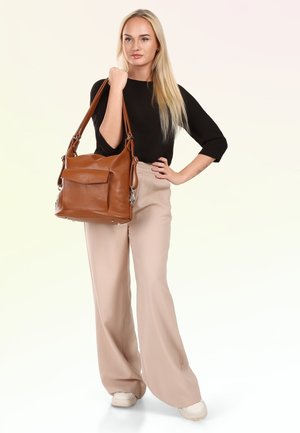 Vrouw met lang blond haar, een zwarte top en beige wijde broek, die een grote bruine leren handtas op de schouder draagt.