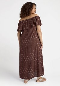 Abito maxi marrone con spalle scoperte, top con volants, motivi a freccia dorati, tessuto fluido, indossato con sandali color beige.