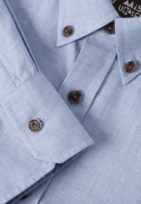 Cămașă bleu deschis, cu butoni, realizată dintr-un material texturat. Prezintă butoane închise la culoare și un mic logo pe manșetă. Design elegant al gulerului.