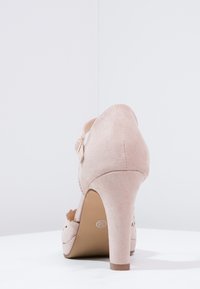 Beżowy zamszowy but na wysokim obcasie z paskiem kostkowym i klamrą, pokazany z tyłu na białej powierzchni.