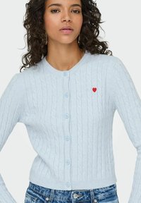 Gaišzils kardigans ar kabeļu adījumu un garām piedurknēm, apaļu kakla izgriezumu, pogu aizdari un mazu sarkanu sirds izšuvumu.