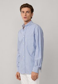 Joven vestido con una camisa de botones a rayas verticales en azul claro y blanco, con un bolsillo en el pecho y pantalones blancos, de pie frente a un fondo liso.