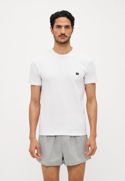 Tricou alb din bumbac cu mâneci scurte, guler rotund și patch cu logo negru pe pieptul stâng. Asortat cu pantaloni scurți gri deschis.