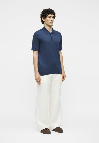 Άνδρας με σγουρά μαλλιά που φοράει μπλε σκούρο κοντομάνικο πόλο, άσπρο παντελόνι με φαρδιά μπατζάκια και καφέ παπούτσια, στέκεται μπροστά σε ένα απλό φωτεινό φόντο.