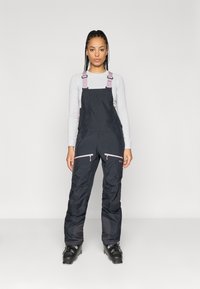 Os macacões pretos à prova d'água apresentam alças rosas ajustáveis, bolsos com fecho de correr e pernas de calças com um acabamento texturizado e elegante. Usados com botas de esqui.